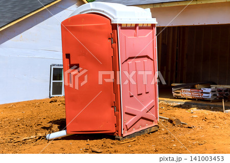 Portable toilet stands alone on dirt ground next to building site with construction materials. Portable toilet stands alone on dirt ground next to building site with construction materials. 141003453