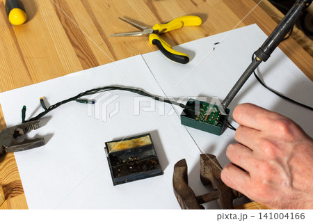 A man works on fixing a broken Christmas tree garland by soldering wires together. He uses tools and parts on a wooden table, focusing on the repair process during the holiday season A man works on fixing a broken Christmas tree garland by soldering wires together. He uses tools and parts on a wooden table, focusing on the repair process during the holiday season 141004166