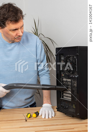 A 53-year-old man cleans dust from a computer system unit with a vacuum cleaner in a tidy room. He carefully removes dirt to keep the device running well A 53-year-old man cleans dust from a computer system unit with a vacuum cleaner in a tidy room. He carefully removes dirt to keep the device running well 141004167