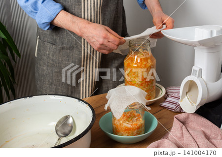 A 53-year-old man prepares to ferment cabbage and carrots in his kitchen. He carefully places a jar filled with vegetables on a bowl. The workspace shows various tools A 53-year-old man prepares to ferment cabbage and carrots in his kitchen. He carefully places a jar filled with vegetables on a bowl. The workspace shows various tools 141004170