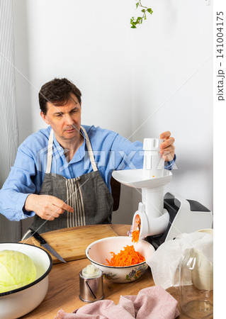 A 53-year-old man is in his kitchen preparing cabbage and carrots for fermentation. He uses a machine to shred the vegetables before placing them in a bowl A 53-year-old man is in his kitchen preparing cabbage and carrots for fermentation. He uses a machine to shred the vegetables before placing them in a bowl 141004175