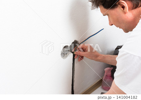 Worker runs new electrical wires into a cutout in a white apartment wall. This task prepares for the installation of an electrical outlet near the baseboard Worker runs new electrical wires into a cutout in a white apartment wall. This task prepares for the installation of an electrical outlet near the baseboard 141004182