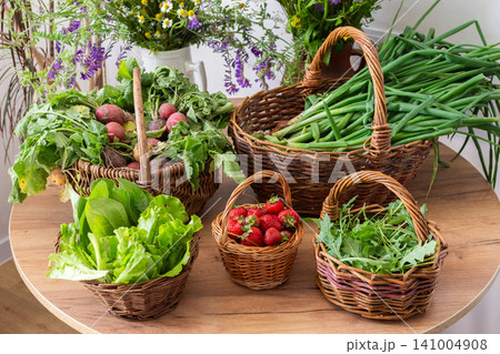 Freshly picked organic vegetables and strawberries are arranged in woven baskets on a wooden table. These local harvests show vibrant greens and red strawberries in a sunny setting Freshly picked organic vegetables and strawberries are arranged in woven baskets on a wooden table. These local harvests show vibrant greens and red strawberries in a sunny setting 141004908