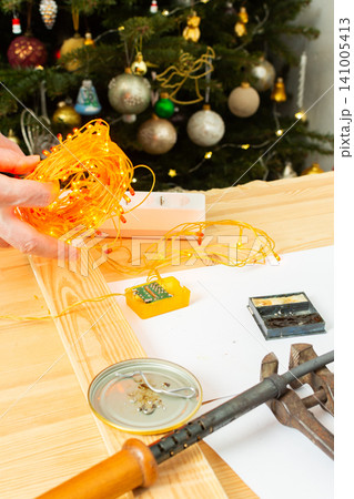 A man fixes a Christmas tree garland and lights on a table at home. He is checking the connection and making adjustments to ensure everything works for the holiday season 141005413