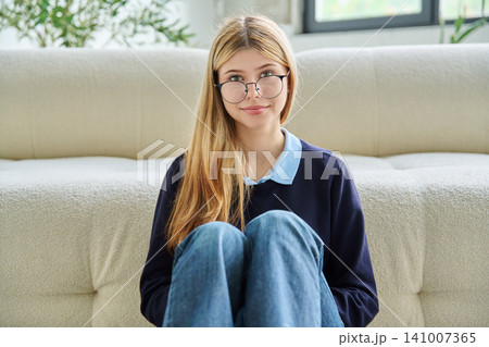 Smiling teenage student girl in glasses sitting on floor hugging knees. 141007365