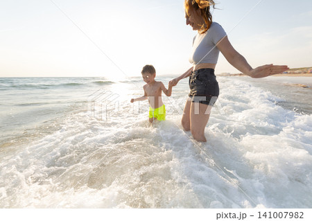 Woman and child play in ocean waves during sunset, with the sun reflecting on the water and beach in the background, creating a vibrant coastal scene 141007982