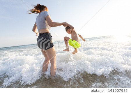 Woman in gray shirt and shorts holds boy's hand as he jumps into ocean waves during sunny day at beach, water splashing around them Woman in gray shirt and shorts holds boy's hand as he jumps into ocean waves during sunny day at beach, water splashing around them 141007985