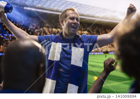 Football supporters shouting with raised arms in sports arena during soccer match day. Passionate crowd in live sports venue creating fan excitement and stadium atmosphere for favorite team 141009894