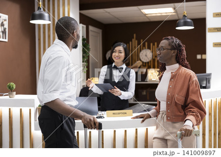 Hotel concierge welcoming travelers couple at reception counter of resort lobby. Guests with luggage complete the registration and check-in process while hotel staff provides customer service. Hotel concierge welcoming travelers couple at reception counter of resort lobby. Guests with luggage complete the registration and check-in process while hotel staff provides customer service. 141009897