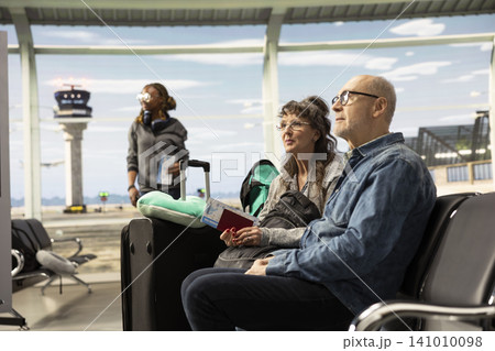 Smiling elderly man and woman sitting in airport lounge with luggage, having a chat and planning their trip while waiting for their plane. Retirees embracing freedom and bucket list travel goals. 141010098