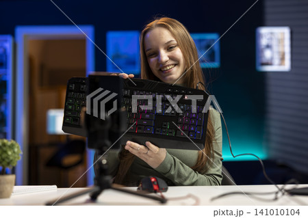 Young woman vlogger testing gaming keyboard, showing buttons and accessory compatibility. Content creator filming a sponsored review video, sharing advice while promoting product. Young woman vlogger testing gaming keyboard, showing buttons and accessory compatibility. Content creator filming a sponsored review video, sharing advice while promoting product. 141010104
