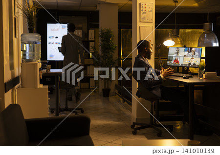 African american male, online videocall, office desk, working late after hours, man focused on monitor to lead briefing presentation and engagement with colleagues across time zones through chat. African american male, online videocall, office desk, working late after hours, man focused on monitor to lead briefing presentation and engagement with colleagues across time zones through chat. 141010139