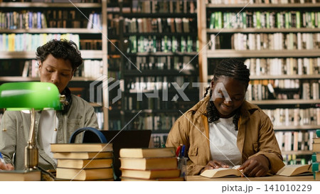 Black woman student reading open book at shared library table, study area with soft focus on shelves in quiet space. Lecture in study session in academic workspace. 141010229