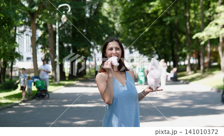 A woman enjoys eating cotton candy in a relaxed and pleasant park pathway surrounded by trees and people, capturing a summer fun family outing with vibrant blue and lush. 141010372