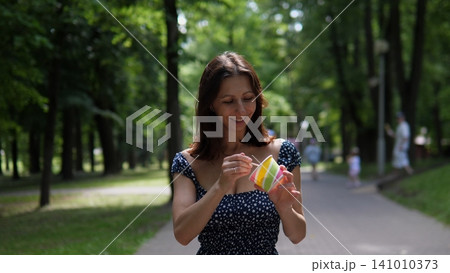 Woman eating ice cream, in park, with pathway, trees, people in the background, with paper cup with colorful, wooden stick for ice texture, from a frontal view perspective. 141010373