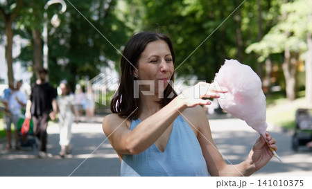 Woman holding cotton candy, in outdoor park setting with trees and people in, with cotton candy, people walking, trees, with cotton candy on stick texture, and summer mood. Woman holding cotton candy, in outdoor park setting with trees and people in, with cotton candy, people walking, trees, with cotton candy on stick texture, and summer mood. 141010375