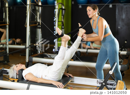 Girl trainer helps mature woman to perform exercises in lying position on pilates reformer Girl trainer helps mature woman to perform exercises in lying position on pilates reformer 141012050