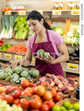 Adult woman seller puts artichokes in vegetable shop 141012099