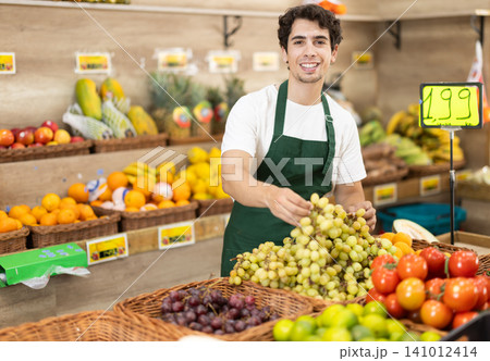Male seller wearing apron holding grape in hand in grocery shop 141012414
