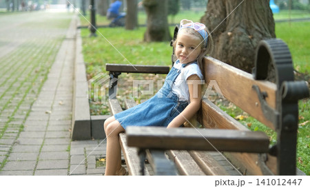 Small happy child girl sitting on a bench resting in summer park. Small happy child girl sitting on a bench resting in summer park. 141012447