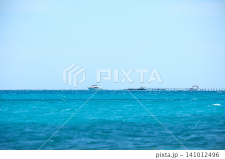 Seascape with ripple surface of blue sea water with distant ship at the end of long pier floating on calm waves. Seascape with ripple surface of blue sea water with distant ship at the end of long pier floating on calm waves. 141012496