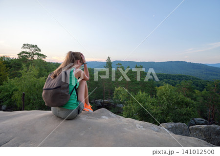 Young sad woman hiker sitting alone on rocky mountain on warm evening. Solitude and resting from everyday rush concept Young sad woman hiker sitting alone on rocky mountain on warm evening. Solitude and resting from everyday rush concept 141012500