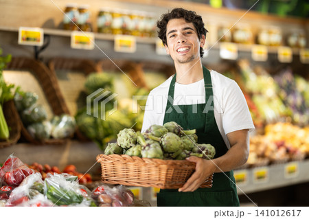 Male seller wearing apron holding artichokes in hand in grocery shop Male seller wearing apron holding artichokes in hand in grocery shop 141012617