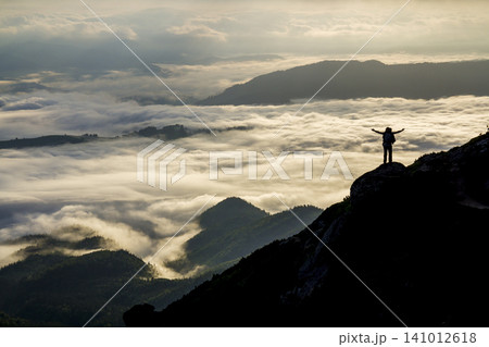 Wide mountain panorama. Small silhouette of tourist with backpack on rocky mountain slope with raised hands over valley covered with white puffy clouds. Beauty of nature, tourism and traveling concept Wide mountain panorama. Small silhouette of tourist with backpack on rocky mountain slope with raised hands over valley covered with white puffy clouds. Beauty of nature, tourism and traveling concept 141012618