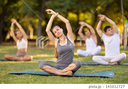 Woman stretching in lotus position during group yoga in park Woman stretching in lotus position during group yoga in park 141012727
