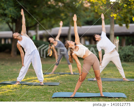 Yoga lesson visitors with coach perform Trikonasana in park 141012775