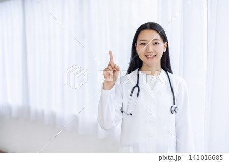 明るい窓辺で人差し指を立ててカメラ目線で微笑む若い日本人の女性医師 明るい窓辺で人差し指を立ててカメラ目線で微笑む若い日本人の女性医師 141016685