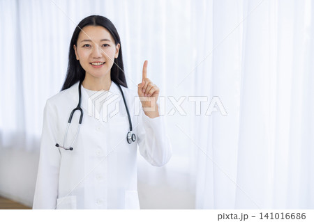 明るい窓辺で人差し指を立ててカメラ目線で微笑む若い日本人の女性医師 明るい窓辺で人差し指を立ててカメラ目線で微笑む若い日本人の女性医師 141016686