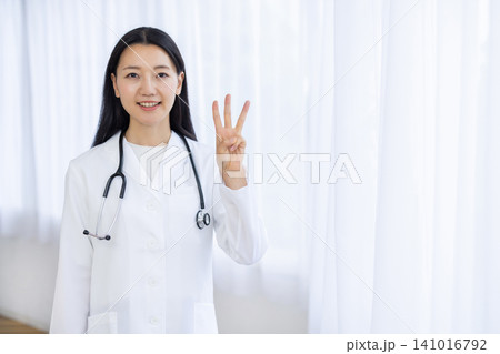 明るい窓辺で指を3本たててカメラ目線で微笑む若い日本人の女性医師 141016792