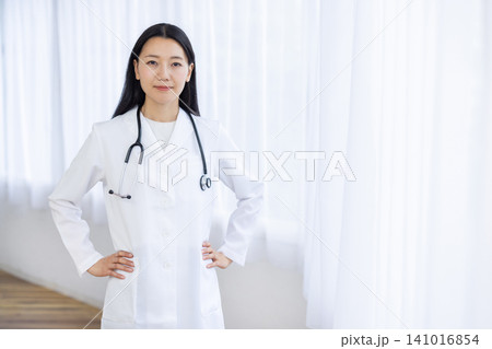 明るい窓辺で腰に手を当ててカメラ目線で微笑む若い日本人の女性医師 明るい窓辺で腰に手を当ててカメラ目線で微笑む若い日本人の女性医師 141016854