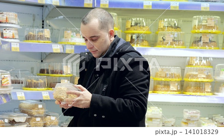 Male shopper browsing packaged desserts in refrigerated bakery section, intently comparing cake varieties and contemplating sweet purchase options 141018329