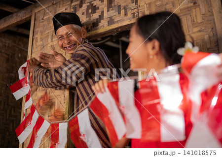Smiling elderly man waving beside blurred woman holding red and white flags inside rustic woven wooden house with warm natural light, cultural celebration, joyful community moment Smiling elderly man waving beside blurred woman holding red and white flags inside rustic woven wooden house with warm natural light, cultural celebration, joyful community moment 141018575