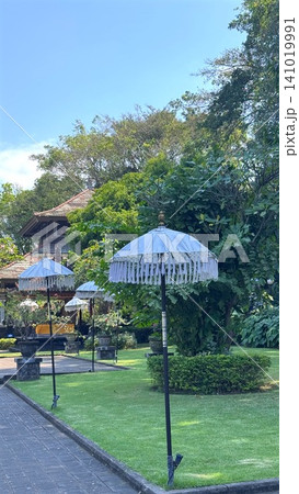 serene bali temple garden with traditional umbrellas and lush greenery on a brick walkway, peaceful setting with potted plants and wooden structures, editorial visual resource for travel. 141019991