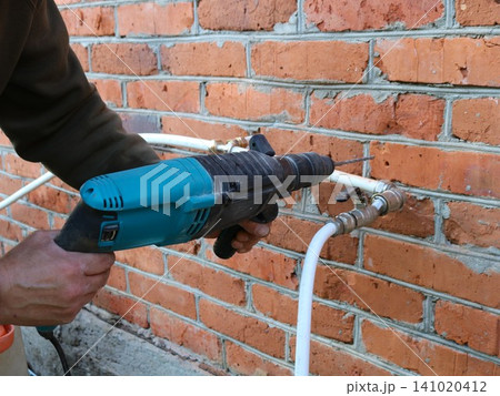 A man is drilling into a red brick wall using a powerful turquoise rotary hammer. White water pipes with metal valves and fittings are neatly laid out next to the drill bit. The process of installing A man is drilling into a red brick wall using a powerful turquoise rotary hammer. White water pipes with metal valves and fittings are neatly laid out next to the drill bit. The process of installing 141020412