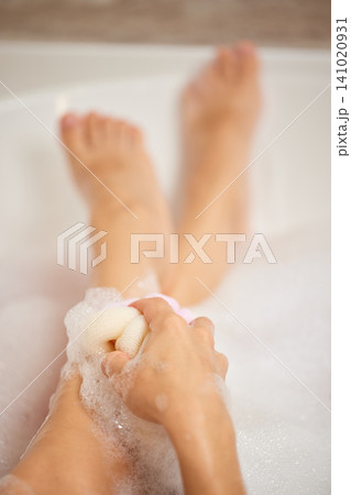 Woman scrubbing her legs with a sponge, creating soap bubbles while relaxing in a warm bathtub for personal hygiene and self-care Woman scrubbing her legs with a sponge, creating soap bubbles while relaxing in a warm bathtub for personal hygiene and self-care 141020931