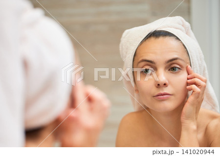 Young woman with towel on head touching face wrinkles looking at reflection in bathroom mirror, analyzing skin aging Young woman with towel on head touching face wrinkles looking at reflection in bathroom mirror, analyzing skin aging 141020944
