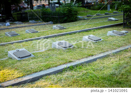 Rows of flat grave markers on grassy cemetery grounds with soft sunlight, trimmed hedges, stone pathways, and calm memorial setting across maintained lawn, with grave, marker, cemetery, memorial 141021091