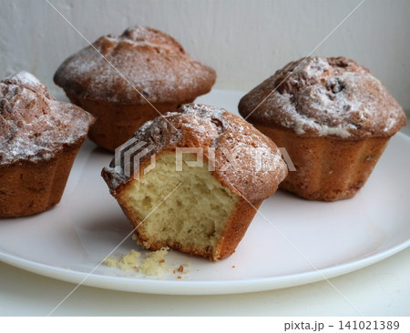 Four golden-brown homemade muffins, dusted with powdered sugar, are arranged on a white plate. One of the muffins has a bite taken out of it, revealing a fluffy, airy crumb and a few crumbs. 141021389