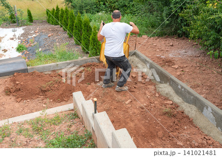 A construction worker in a white t-shirt pushing a yellow wheelbarrow with soil during backyard renovation and landscaping works near a new concrete retaining wall. High-angle view of a builder A construction worker in a white t-shirt pushing a yellow wheelbarrow with soil during backyard renovation and landscaping works near a new concrete retaining wall. High-angle view of a builder 141021481