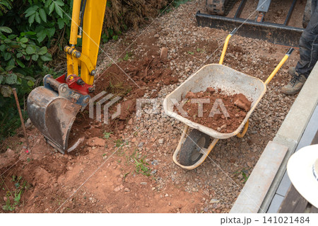 An excavator bucket is actively loading soil into a wheelbarrow from a construction site, viewed from a top-down perspective. The scene is set in a commercial environment with natural light. An excavator bucket is actively loading soil into a wheelbarrow from a construction site, viewed from a top-down perspective. The scene is set in a commercial environment with natural light. 141021484