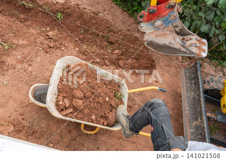 Top-down close-up shot of a mini excavator bucket loading fresh brown soil into a metal wheelbarrow on a construction site. High-quality image for earthwork services. Mini digger bucket filling a Top-down close-up shot of a mini excavator bucket loading fresh brown soil into a metal wheelbarrow on a construction site. High-quality image for earthwork services. Mini digger bucket filling a 141021508