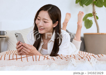 ベッドに寝転んでスマートフォンを見る笑顔の若い女性 141021829