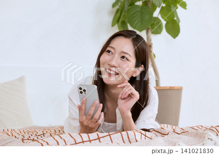 ベッドに寝転びスマートフォンを持ち微笑む若い女性 141021830