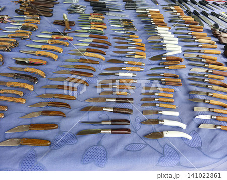XALO, SPAIN - JUNE 20, 2026.Numerous folding pocket knives with different wooden and plastic handles are laid out on a blue table at a street market. People explore the collection and shop. XALO, SPAIN - JUNE 20, 2026.Numerous folding pocket knives with different wooden and plastic handles are laid out on a blue table at a street market. People explore the collection and shop. 141022861