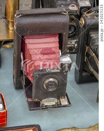 XALO, SPAIN - JUNE 20, 2026.A rare antique folding bellows camera with a red leather body is set on display at a flea market. Surrounding it are various vintage electronics for sale. Shoppers browse t XALO, SPAIN - JUNE 20, 2026.A rare antique folding bellows camera with a red leather body is set on display at a flea market. Surrounding it are various vintage electronics for sale. Shoppers browse t 141023133