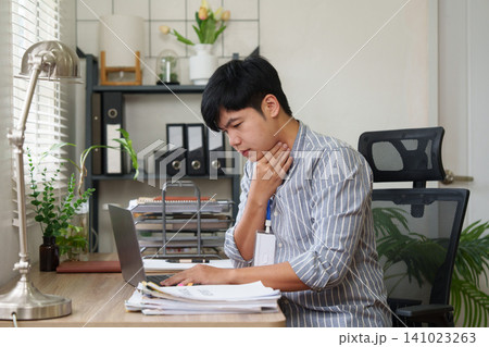 Asian businessman with sore throat touching neck while working on laptop in office Asian businessman with sore throat touching neck while working on laptop in office 141023263